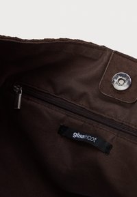 Vnitřek hnědé textilní tašky s kovovým patentním knoflíkem, kapsou na zip a černou etiketou s bílým nápisem "ginatricot."