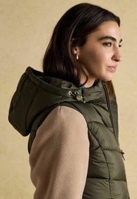 Oliwkowa kamizelka puchowa z kapturem, z zamkiem z przodu i złotymi elementami, zestawiona z beżową polarową bluzą z długim rękawem.
