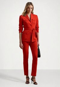 Rødt skreddersydd dress med belte i midjen, to brystlommer og smale bukser. Brukt med svarte sandaler og en liten håndveske. Glatt tekstur.