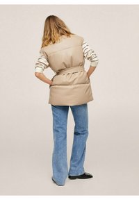 Beige sleeveless puffer vest with belt, textured finish, worn over a striped long-sleeve shirt, paired with flared blue jeans and black shoes.