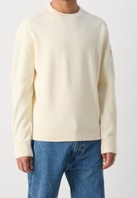 Světlý krémový svetr s žebrovaným lemem a manžetami, s kapsou na zip na levém rukávu a hladkou, texturovanou tkaninou.