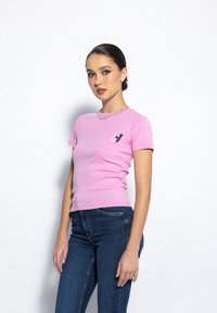 Różowa bawełniana koszulka z krótkim rękawem, posiadająca małe czarne logo na lewej stronie klatki piersiowej. Noszona z ciemnoniebieskimi dżinsami. Gładka tekstura.