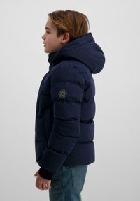 Jongen in een marineblauwe donsjack met capuchon, zijaanzicht tegen een effen achtergrond, kijkt iets naar rechts, hand rustend aan zijn zijde.