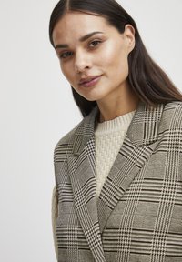 Blazer à carreaux en noir et crème, avec un col structuré. Porté par-dessus un pull beige texturé, mettant en valeur le look superposé.
