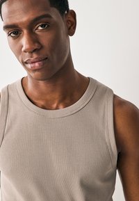 Débardeur beige côtelé avec un col rond, présentant une coupe ajustée et une texture lisse. Coutures visibles et accents aux épaules.