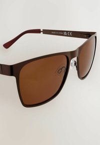 Braune Sonnenbrille mit rechteckiger Form, ausgestattet mit dunkelbraunen Gläsern und einem schlanken Kunststoffrahmen. Enthält transparente Nasenpolster und rote Bügel.