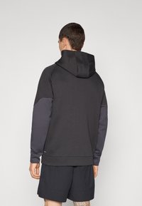 Hættebluse i mørkegrå, med en kontrasterende lysere grå panel på ærmerne, blød tekstur og ribbet kant. Bæres med sorte shorts.