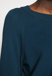 Maglione a maglia verde scuro, con scollatura rotonda, polsini a coste e una struttura liscia. Dettagli dei punti visibili alla cucitura della spalla.