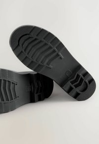 Svart gummisula med texturerat grepp, med räfflor och mönstrade sektioner för bättre fäste. Designen är platt med ett rundat tåområde.