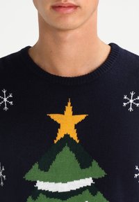 Pull en tricot bleu marine avec un motif d'arbre de Noël, une étoile jaune en haut et des flocons de neige blancs sur les épaules.