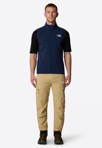 Navy sleeveless vest over a black short-sleeve shirt, paired with beige cargo pants featuring side pockets. Black hiking boots complete the outfit.