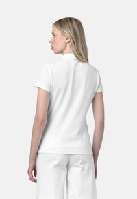 Maglietta polo bianca con maniche corte e colletto, realizzata in cotone, con una superficie liscia. Indossata con pantaloni bianchi, vista da dietro.