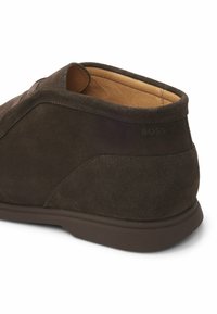 Brown suede ankle boot featuring a rounded shape, contrasting tan lining, and subtle side branding. Rubber sole with a textured finish.