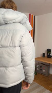 Φουσκωτό, ανοιχτό γκρίζο μπουφάν με μεγάλο καπέλο και καπιτονέ υφή. Διαθέτει κρυμμένα φερμουάρ και χαλαρή εφαρμογή. Το φόντο περιλαμβάνει πολύχρωμες ρίγες.