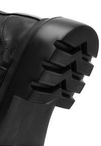 Bota de cuero negro con una suela de goma gruesa y texturizada que presenta patrones en zigzag para tracción. Parte superior lisa con un diseño de punta redonda.
