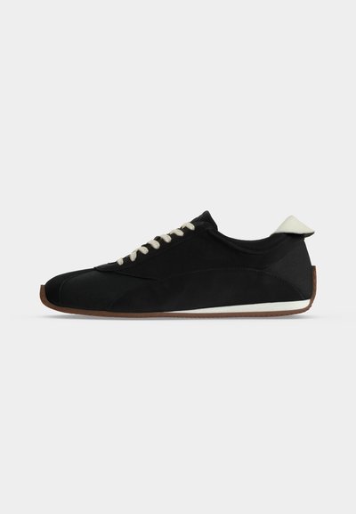 Μαύρο sneaker χαμηλής κοπής με λευκά κορδόνια, λευκή γλωττίδα στη φτέρνα και καφέ ελαστική σόλα σε απλό ανοιχτό φόντο.