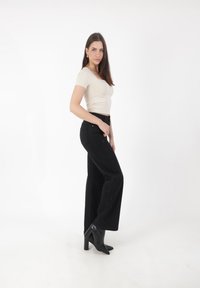 Top aderente beige, jeans neri a gamba larga con vita alta e leggera trama, abbinati a tacchi a punta neri, su sfondo bianco.