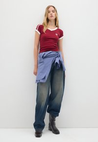 Top corto rojo con manga corta y detalles blancos, combinado con unos vaqueros azules de gran tamaño y una camisa de cuadros azul atada a la cintura. Botines negros.