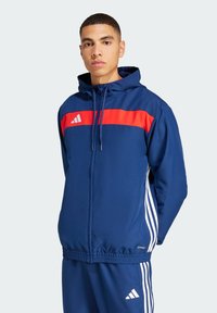 Marine atletisk jakke med rød vandret stribe, lynlås foran, justerbar hætte og hvide tre-striber detaljer på ærmerne. Glat stof.