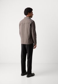 Chaqueta gris de botones con cuello, corte relajado y mangas largas. Pantalones y zapatos negros completan el conjunto, visto desde la parte de atrás.