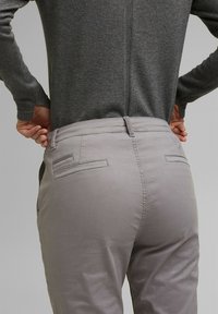 Personne portant un pantalon gris clair et une chemise à manches longues gris foncé, ajustant la taille du pantalon par l'arrière.