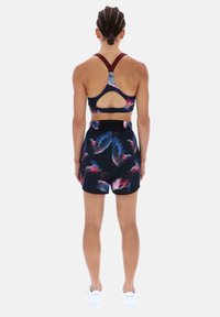 Sujetador deportivo con tirantes burdeos y espalda con recorte; combinado con pantalones cortos negros con un patrón abstracto colorido. Zapatillas blancas completan el conjunto.