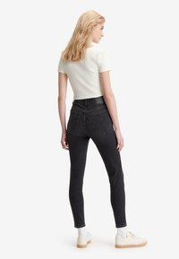 Fiatal nő, aki szőke hajú, háttal áll, fehér bordázott rövid ujjú pólót, fekete farmert, fehér zoknit és krémszínű sportcipőt visel.