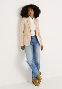 Blazer rose clair cintré porté sur une chemise blanche à volants, associé à un jean droit bleu et des bottines beiges, présenté sur un fond neutre.