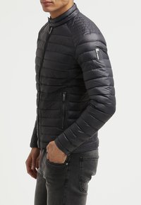 Veste matelassée bleu marine avec col montant, texture matelassée, fermeture éclair à l'avant et poches, présentant des coutures horizontales et un petit logo sur la manche.