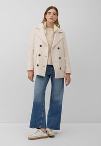 Frau in einem beigen doppelreiherigen Mantel, hellem beigen Pullover, blauen weit geschnittenen Jeans und weißen Sneakers, vor einem einfarbigen Hintergrund stehend.