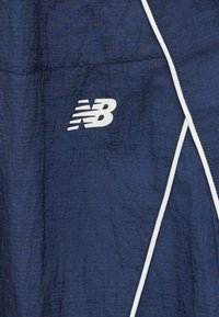 Σκούρο μπλε υφασμάτινο σακάκι με λευκές λεπτομέρειες περιγράμματος και λευκό λογότυπο New Balance στο μπροστινό μέρος.
