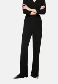 Black ribbed pants with a flared design, featuring a textured fabric and a high waistband. Worn with black shoes, accented with gold tips.