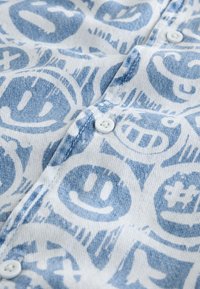 Jeanshemd mit einem hellblauen und weißen Muster verschiedener Smiley-Gesichter. Der Stoff hat eine weiche Textur, weiße Knöpfe sind sichtbar.