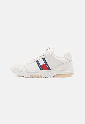 Zapatilla blanca con parte superior de piel suave, que presenta un acento a rayas azul y roja en el costado, suela texturizada y cordones redondos.