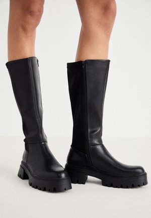 Bottes noires montantes jusqu'aux genoux en cuir lisse, avec une fermeture éclair latérale et une semelle en caoutchouc épaisse à crampons texturés.