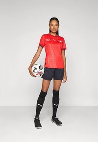 Rødt fotballtrøye med mønster, svarte shorts, svarte knelange sokker og svarte fotballsko. Spilleren holder en hvit fotball med design.