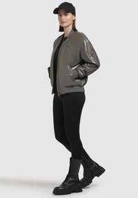 Glanzende grijze bomberjack met geribde boorden, gecombineerd met een zwart top en legging. Zwarte veter combat boots maken de outfit af. Zwarte pet gedragen.