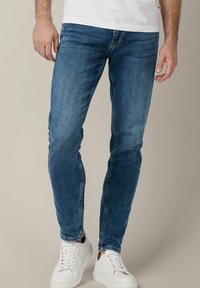 Slim-fit niebieskie dżinsy z wyblakłym wykończeniem, pięć kieszeni i subtelne detale szycia. W zestawie z białymi sneakersami.