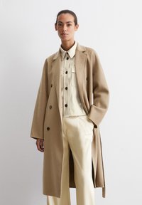 Cappotto in misto lana beige con design doppiopetto, rever a scopertura e grandi bottoni, indossato sopra una camicia button-up crema e pantaloni.