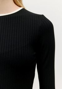 Zwarte geribde lange mouw top met een ronde hals en een contrasterende witte kraagaccent. Zacht, gemarmerd materiaal benadrukt het ontwerp.