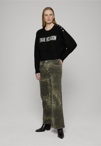 Schwarzer gestrickter Pullover mit dem Schriftzug "TRUE RELIGION" und metallischen Knöpfen auf der Schulter, kombiniert mit khakifarbenen, weit geschnittenen Jeans mit Leopardenmuster und schwarzen Stiefeln.