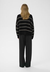 Suéter a rayas negro y beige, de corte oversized, con textura de tejido, combinado con pantalones negros holgados, mostrado desde atrás. Fondo blanco liso.