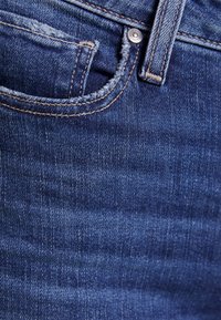 Tummansiniset farkut, joissa kulunut rakenne, pieni etutasku, kontrastipistely ja metallinen nappidetalji.