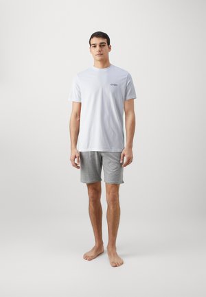 T-shirt bianca in cotone con logo "GUESS" sul lato sinistro del petto, abbinata a pantaloni corti grigi in jersey. Il modello è in piedi a piedi nudi contro uno sfondo semplice.