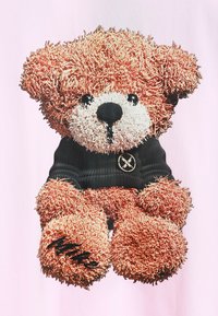 Oso de peluche con pelaje texturizado y esponjoso en color naranja, ojos de botón negros y un suéter negro con un logo. El fondo es rosa.