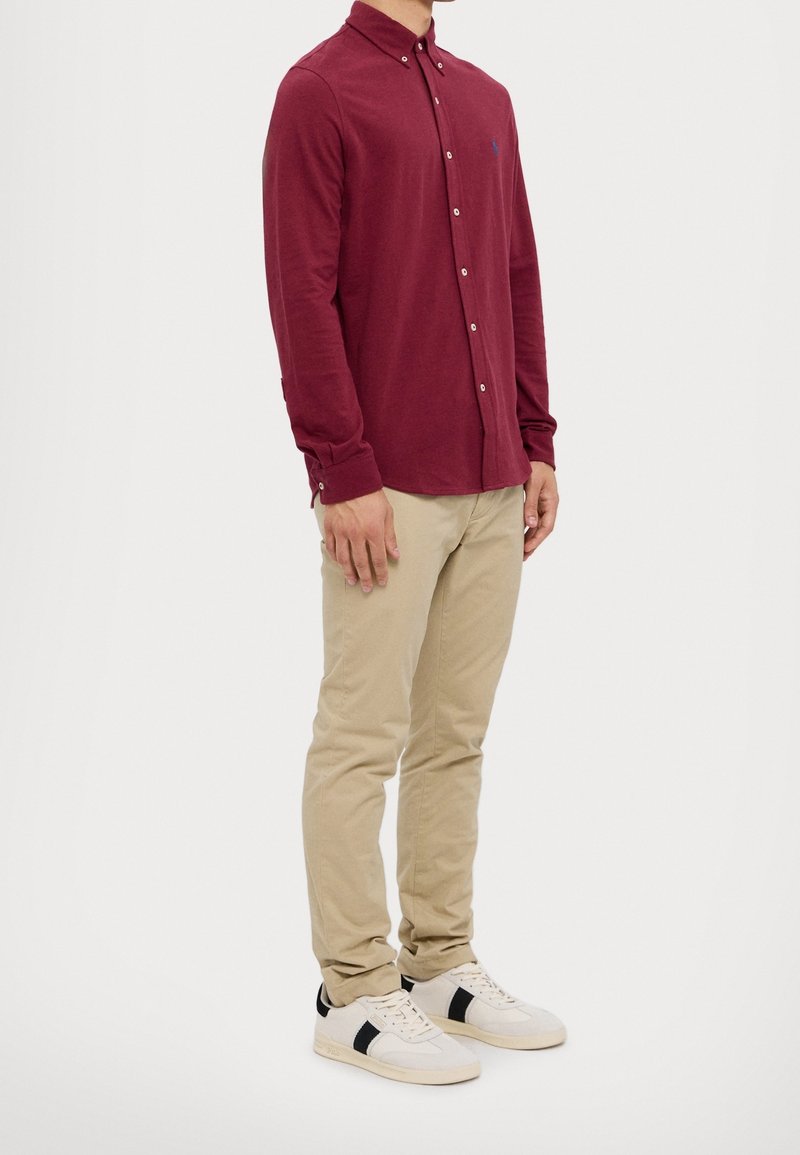 Camisa de manga larga burdeos con botones, pantalones chinos color beige y zapatillas blancas con detalles en negro. Diseño sencillo, corte ajustado y material de algodón.