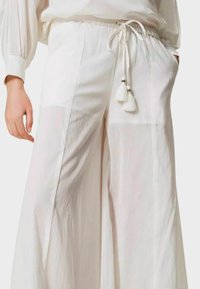Pantaloni bianchi larghi con gamba ampia, vita con cordoncino annodato con pompon, indossati con una maglietta bianca a maniche lunghe abbinata, mano in tasca.