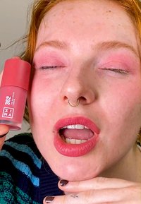 Blush cremoso rosa in un contenitore cilindrico con finitura opaca. Il modello mostra un trucco rosa sugli occhi e sulle labbra, evidenziando un look coordinato nei colori.