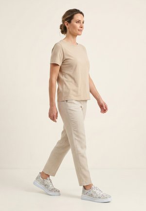 Woman walking, wearing beige short-sleeve shirt, light beige pants, and white floral-patterned sneakers against plain light background.