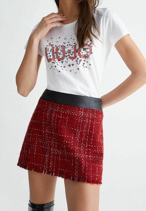 Kobieta nosząca białą koszulkę z czerwonymi wzorami serc i tekstem, w połączeniu z czerwoną spódnicą tweedową z czarnym pasem w talii.